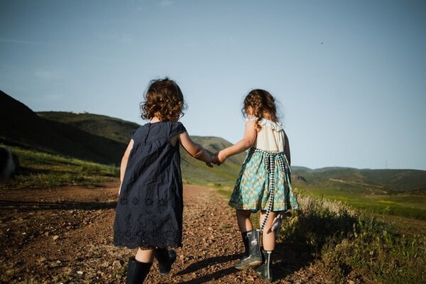 Two preschool aged children in dresses, holding hands walking together on a path through an open field.
