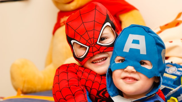Two smiling preschool aged kids, one dressed in a red Spiderman costume and the other dressed in a blue Captain America costume with an A on his mask.