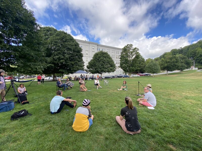Community members are gathered in a circle on the VT State House Lawn. The lawn is green, bordered by green trees, blue sky and white fluffy clouds. Participants are seated on the grass and in lawn chairs.