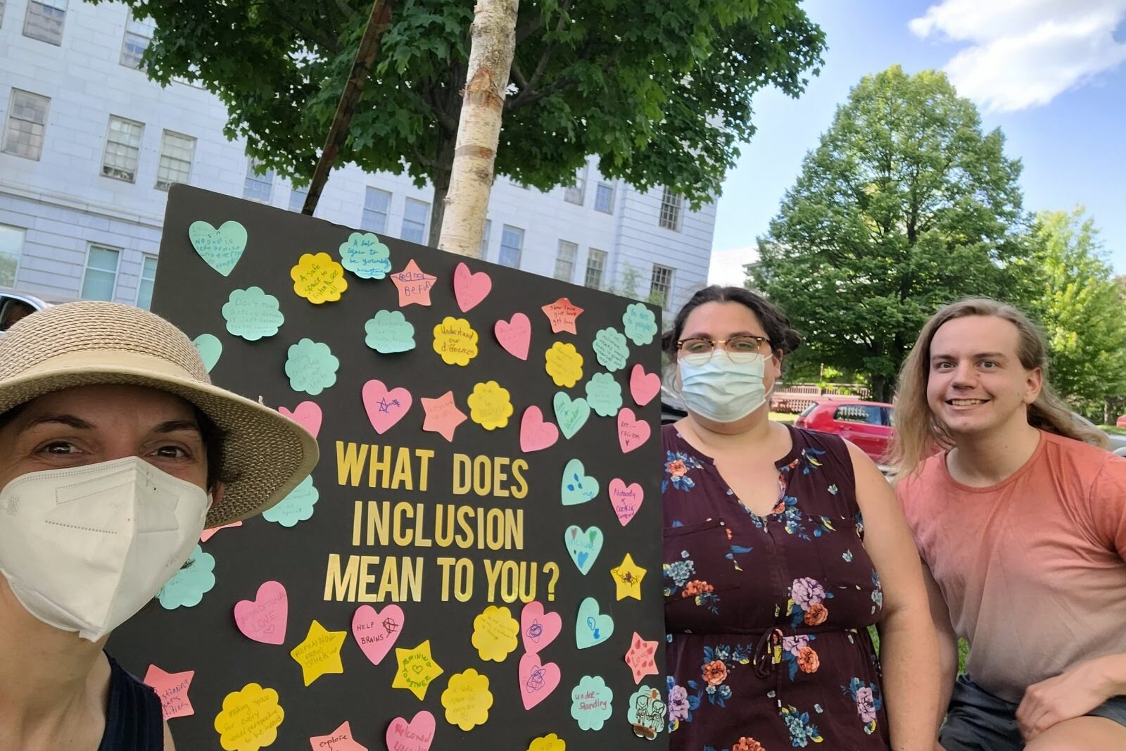 Mel. Sierra and Clem pose in front of a black board reading in gold letters "What Does Inclusion mean to you?" post-its in multiple colors and shapes contain community member responses. They are wearing well-fitting masks. they are smiling.