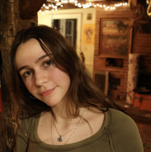 Reagan has long brown hair and is wearing an olive shirt. She is looking at the camera.