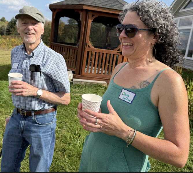Two adults smile on a summer day. One person wears a hat and holds a cup. The other wears sunglasses and holds a cup. They are connecting with a community.